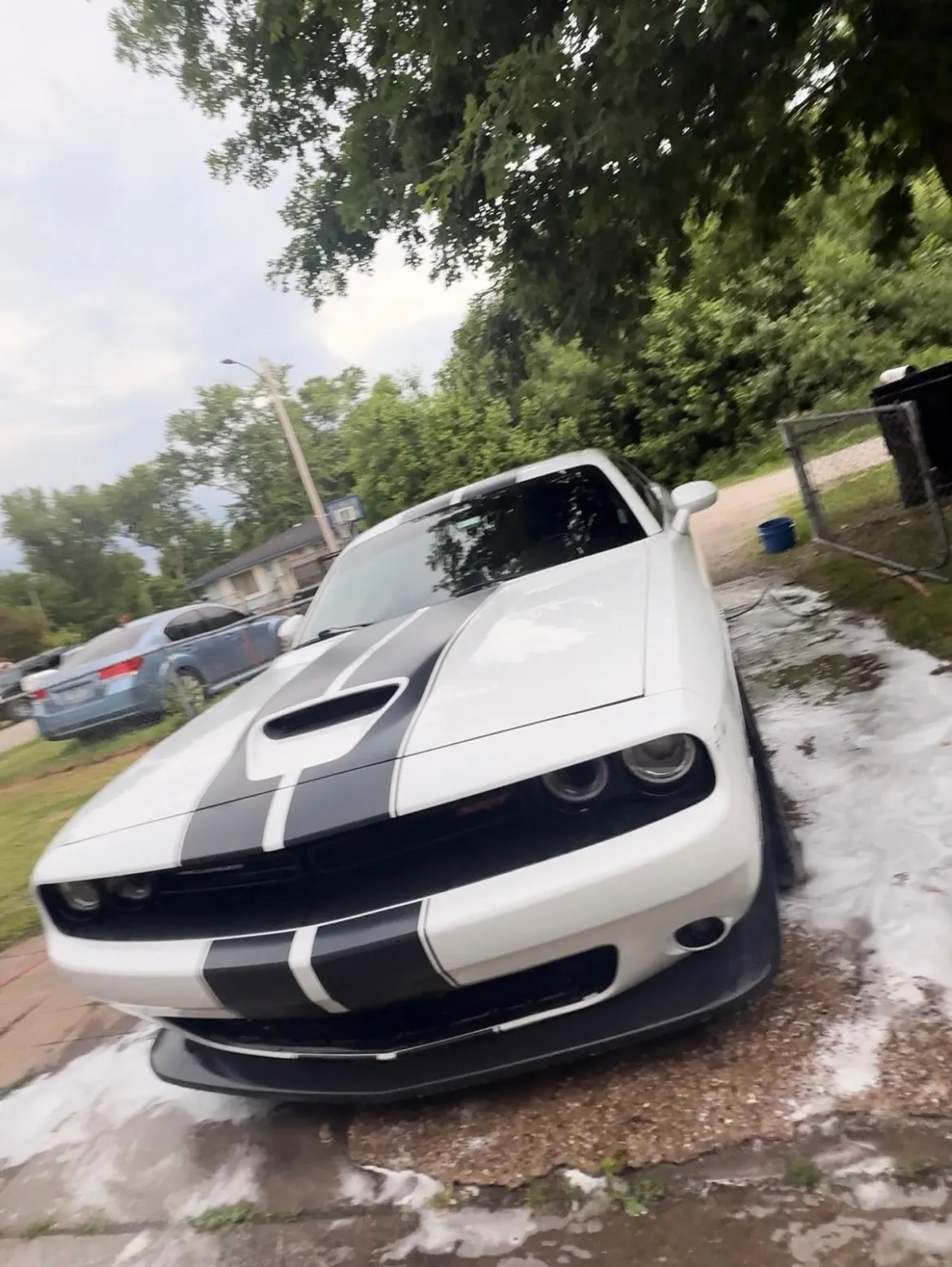 A white Dodge Challenger with racing stripes during an exterior wash, foam runoff visible on the driveway.