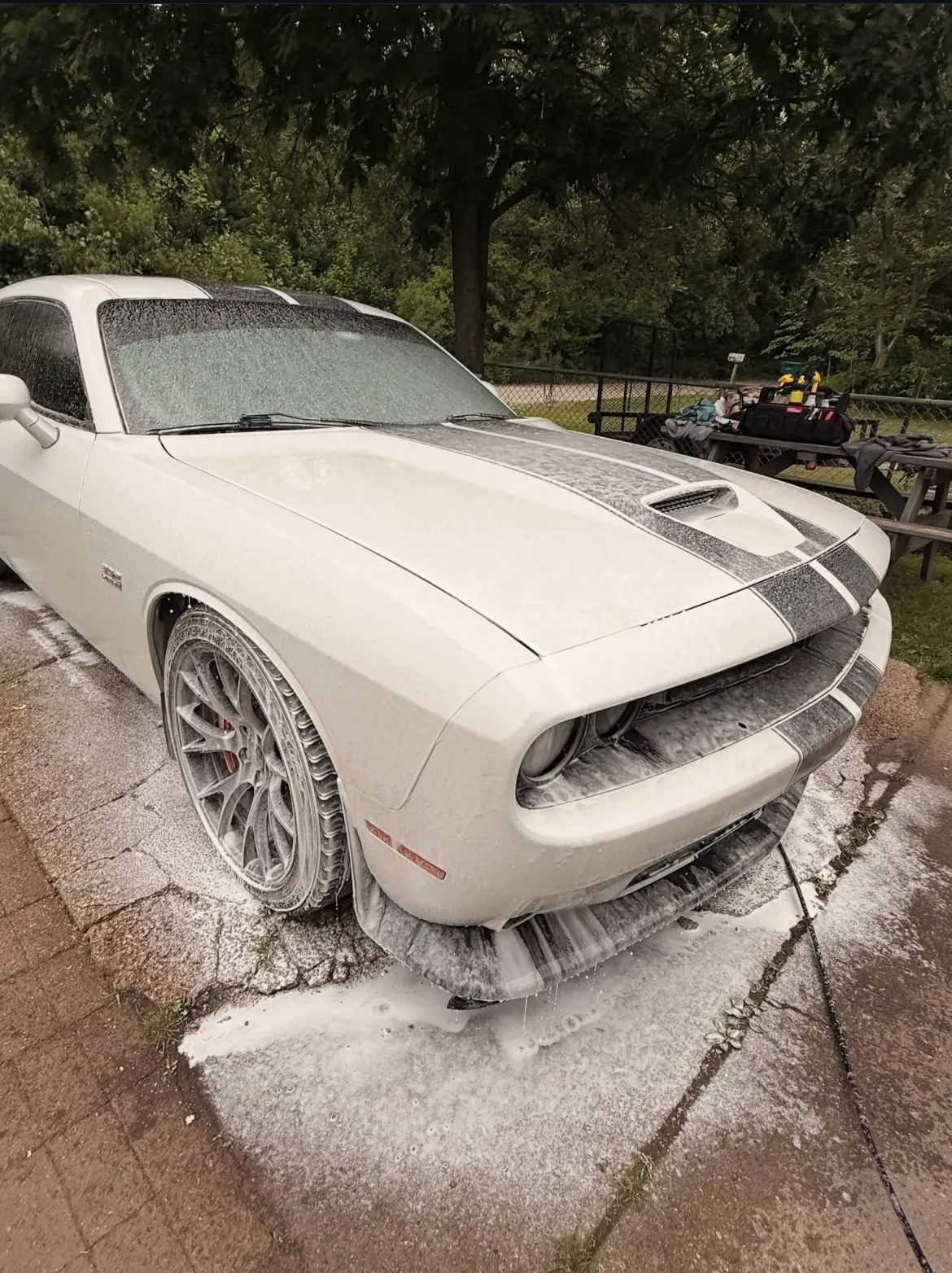 A white Dodge Challenger Scat Pack covered in thick snow foam on a driveway, with detailing equipment set up behind it.